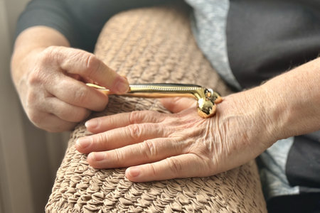 An unknown elderly woman sitting in an armchair makes herself a hand massage with her hand with a massager in the form of two spinning golden rollers. Health maintenance and Promotion Programの写真素材