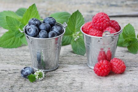 Two small metal bucket filled with ripe raspberries and blueberries, on a background of the old...