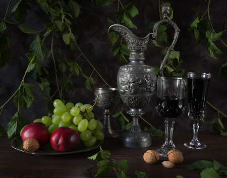 Still life in medieval style with antique pitcher and a goblets filled with red wine as well as walnuts, green grapes and nectarines on a dark background, covered with green plantsの写真素材