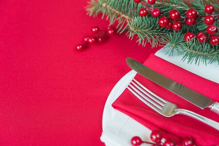 Silver knife and fork lie on the red linen napkin, as well as red holly berries and green spruce branch, which is located on a table covered with a red tablecloth, with space for textの写真素材