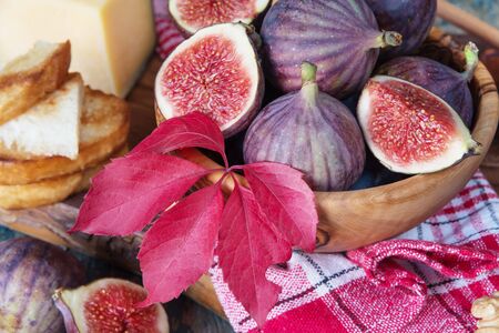 Ripe figs in a wooden bowl, cheese, cane sugar, honey, toast and a checkered napkin on old cutting board as well as red autumn leaves lie on the old wooden tableの写真素材
