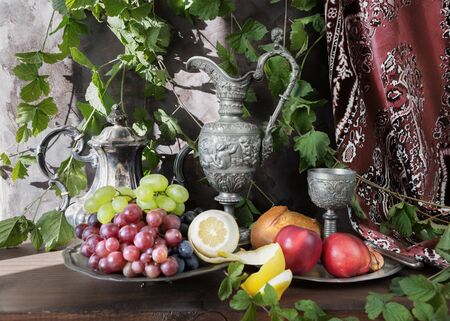 Still life in oriental style with antique pitcher and a goblets filled with wine, grapes, nectarines and lemon on a dark background, covered with green plantsの写真素材