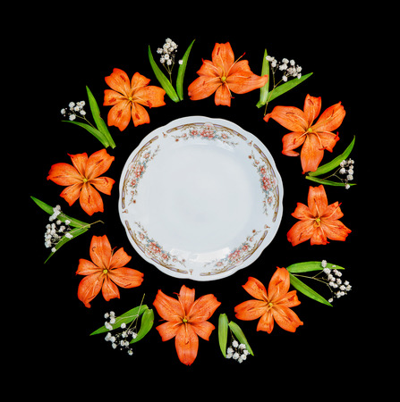 White porcelain empty plate with elegant design, surrounded by beautiful orange flowers lilies arranged in the form of a round frame, isolated on a black backgroundの写真素材
