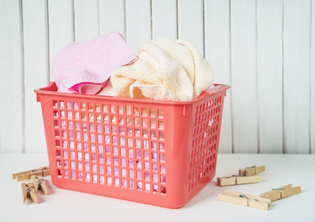 White, beige and pink towels in a red plastic laundry basket and wooden clothespins are on the white wooden backgroundの写真素材