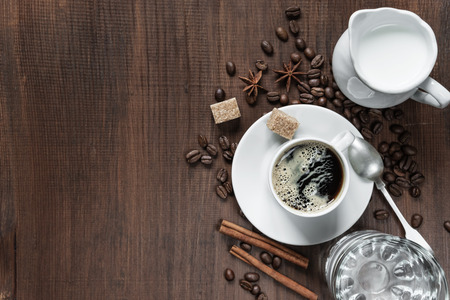 Cup of coffee, cream in a milk jug, glass of water and various spices on the wooden table with...