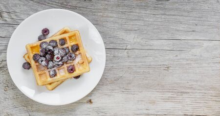 White porcelain plate with sweet waffles and fresh berries, powdered with sugar, on the background of old wooden board, with space for textの写真素材