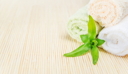 Spa concept: three bath towels twisted into a rolls and green sprout of lucky bamboo with fresh leaves on a beige bamboo mat, with copy-spaceの写真素材