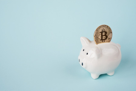 White ceramic piggy bank with golden coin of bitcoin crypto currency, new digital money in cyber world, isolated on a blue backgroundの写真素材
