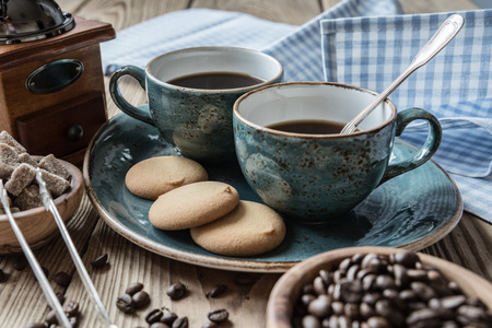 Two blue cups of black coffee, cookies and sugar pieces surrounded by checkered linen cloth and coffee beans on old wooden tableの写真素材