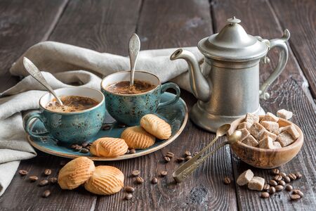 Two blue vintage cups of black coffee, cookies and antique pewter coffee pot surrounded by gray linen napkins, sugar pieces and coffee beans on old wooden tableの写真素材