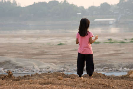 Laotian girl watching the dried out riverbed of Mekong river at Vientiane in Laos. Thailand visible on the opposite side. Shallow depth of field with the girl in focus.の写真素材