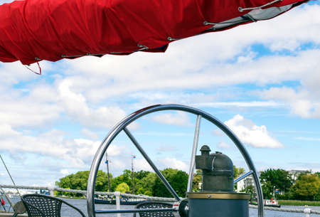 Steering wheel and red sails of tall ship against sky.