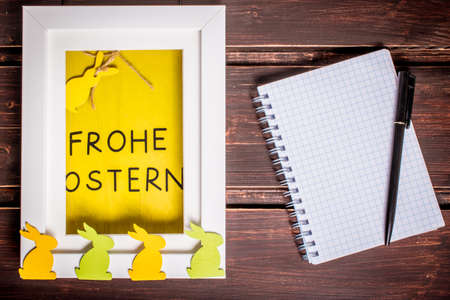 Colorful wooden easter rabbits and yellow happy Easter card in german language "frohe Weihnachten" in the frame. blank notebook with copy space and black pen. template and easter greeting card.の写真素材