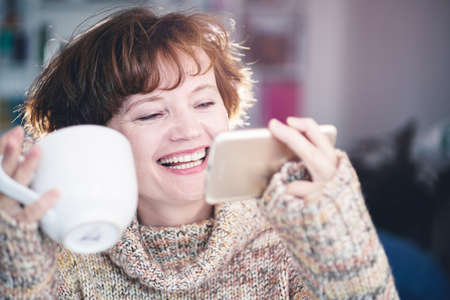 Happy young woman with coffee cup texting on mobile phone. Morning coffee at home. Laughing woman...