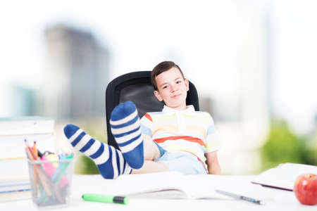 Relaxed casual school boy with legs on desk. cute bored teenager sitting at the desk with school homework. Back to school concept.の写真素材