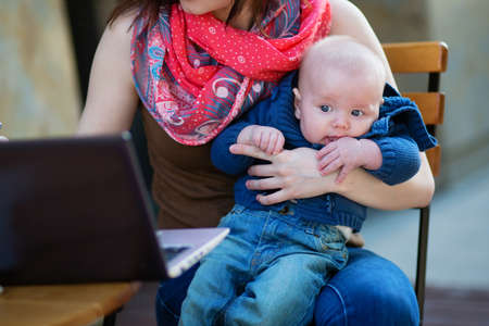 Little 4-month baby in his mother's arms while she's working on her laptopの写真素材