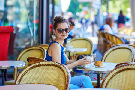 Beautiful young Parisian woman in blue blouse drinking coffee in an outdoor cafe on a summer dayの写真素材