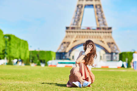 Beautiful young tourist or student girl in Paris sitting on the grass near the Eiffel tower on a summer dayの写真素材