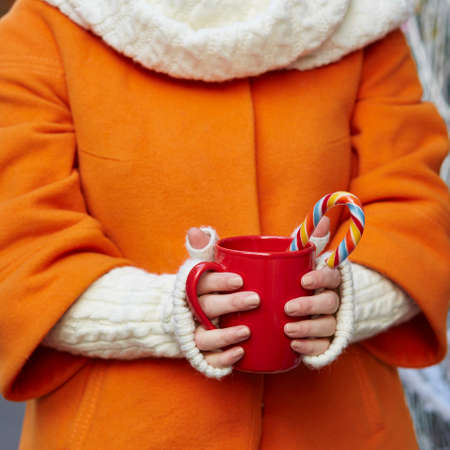 Woman hands in woolen mittens holding a cozy mug with hot cocoa, tea or coffee and a Christmas candy caneの写真素材