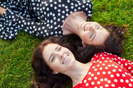 Beautiful twin sisters in red and black polka dot dresses lying on the grass close to each other and smilingの写真素材