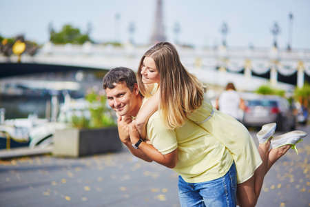 Beautiful young dating couple in Paris near the Alexandre III bridge, Eiffel tower in the background, man is piggybacking his girlfriendの写真素材