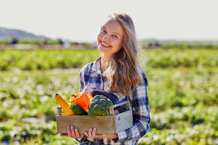 Happy young woman holding wooden crate with fresh organic vegetables from farmの写真素材