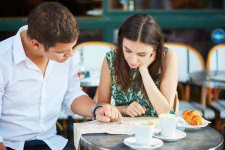Young romantic couple drinking coffee, eating traditional French croissants and planning their itinerary using map in a cozy outdoor cafe in Paris, Franceの写真素材