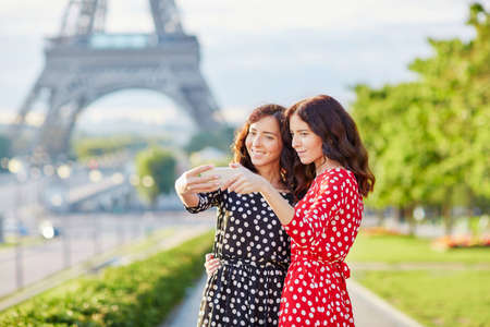 Beautiful twin sisters taking selfie in front of Eiffel Tower while traveling in Paris, France. Happy smiling girls enjoy their vacation in Europeの写真素材