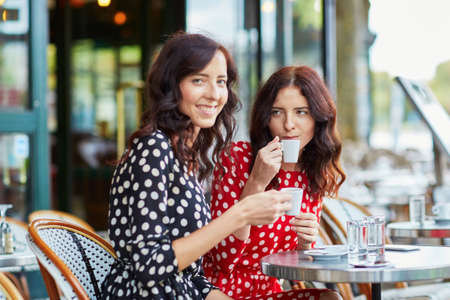 Beautiful twin sisters drinking coffee in a cozy outdoor cafe in Paris, France. Happy smiling girls enjoy their vacation in Europeの写真素材