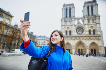 Beautiful young tourist in Paris on a fall day, making selfie with Notre-Dame cathedralの写真素材