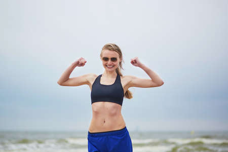 Healthy fitness runner girl wearing sunglasses showing her biceps. Young European woman on beach cardio training taking a rest during workout. Healthy lifestyle and fitness conceptの写真素材