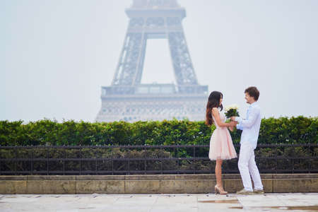 Beautiful romantic couple in love having a date near the Eiffel tower in Paris on a cloudy and foggy rainy day, man is offering huge bunch of white roses to his girlfriendの写真素材