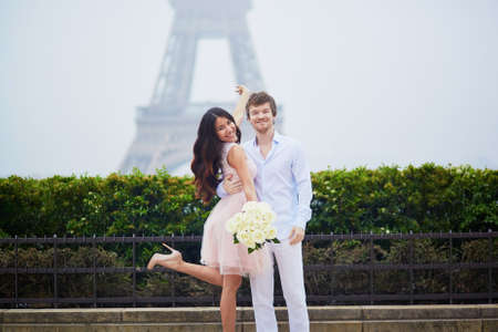 Beautiful romantic couple in love holding bunch of white roses and hugging near the Eiffel tower in Paris on a cloudy and foggy rainy dayの写真素材