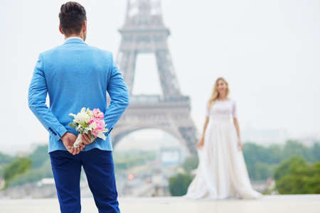 Groom hiding the wedding bouquet behind his back and going to offer it to a bride. Romantic wedding couple near the Eiffel tower in Paris. Surprise proposal conceptの写真素材