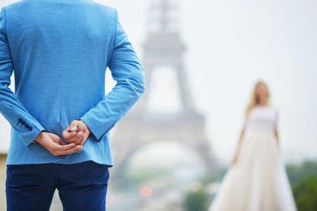Groom hiding the wedding ring behind his back and going to offer it to a bride. Romantic wedding couple near the Eiffel tower in Paris. Surprise proposal conceptの写真素材
