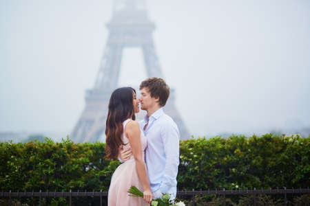 Beautiful romantic couple in love holding bunch of white roses and hugging near the Eiffel tower in Paris on a cloudy and foggy rainy dayの写真素材