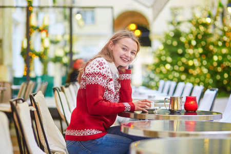 Cheerful young woman drinking hot chocolate in a cozy outdoor Parisian cafe decorated for Christmasの写真素材