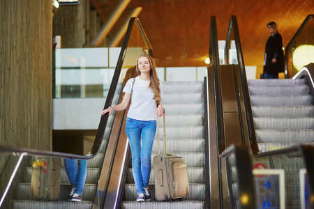 Beautiful young tourist girl with backpack and carry on luggage in international airport, on escalatorの写真素材
