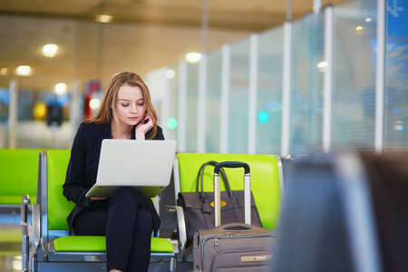 Young elegant business woman with hand luggage in international airport terminal, working on her laptop while waiting for flightの写真素材