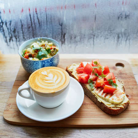 Delicious breakfast, brunch or lunch with hummus and tomato sandwich, salad and fresh hot cappuccino coffee served on cutting board near the window with water drops on wet glass at rainy dayの写真素材