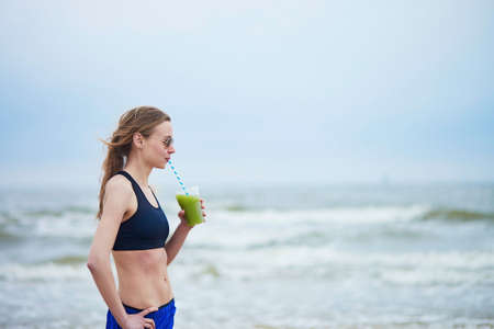 Healthy runner girl drinking green vegetable smoothie from plastic glass on running break. Young woman on beach cardio training taking a rest during workout. Healthy lifestyle, sport and diet conceptの写真素材