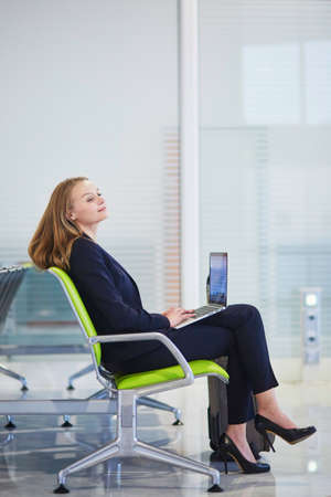 Young elegant business woman with hand luggage in international airport terminal, working on her laptop while waiting for flightの写真素材