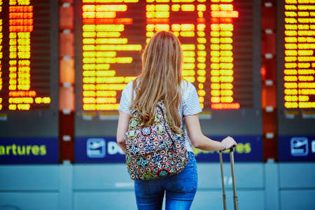 Beautiful young tourist girl with backpack and carry on luggage in international airport, near flight information boardの写真素材