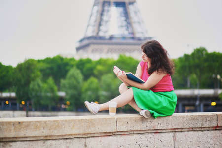 Cheerful young girl with coffee to go reading a book near the Eiffel tower. Student preparing for exam in Paris. International education exchange program in France conceptの写真素材