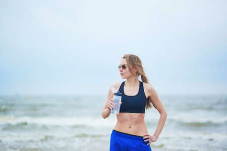 Healthy runner girl drinking water from plastic bottle on running break. Young European woman on beach cardio training taking a rest during workout. Healthy lifestyle, sport and hydration conceptの写真素材
