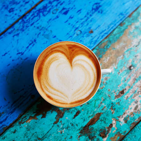 Cup of fresh coffee with heart form milk drawing on blue wooden table, view from above, flat layの写真素材