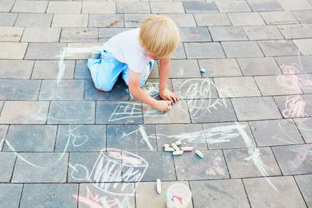 Adorable little boy drawing with colorful chalks on asphalt. Summer activity and creative games for small kidsの写真素材