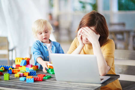 Adorable little boy playing with colorful plastic construction blocks while his mother working on her laptop. Childcare and work conceptの写真素材