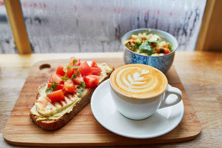 Delicious breakfast, brunch or lunch with hummus and tomato sandwich, salad and fresh hot cappuccino coffee served on cutting board near the window with water drops on wet glass at rainy dayの写真素材