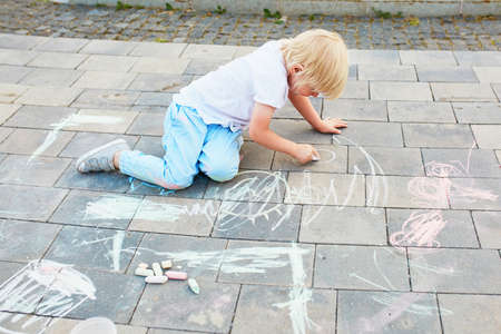 Adorable little boy drawing with colorful chalks on asphalt. Summer activity and creative games for small kidsの写真素材